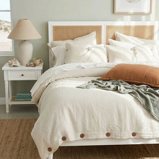 Bedroom with a bed featuring white bedding and brown accents, a nightstand with a lamp, and a window in the background.