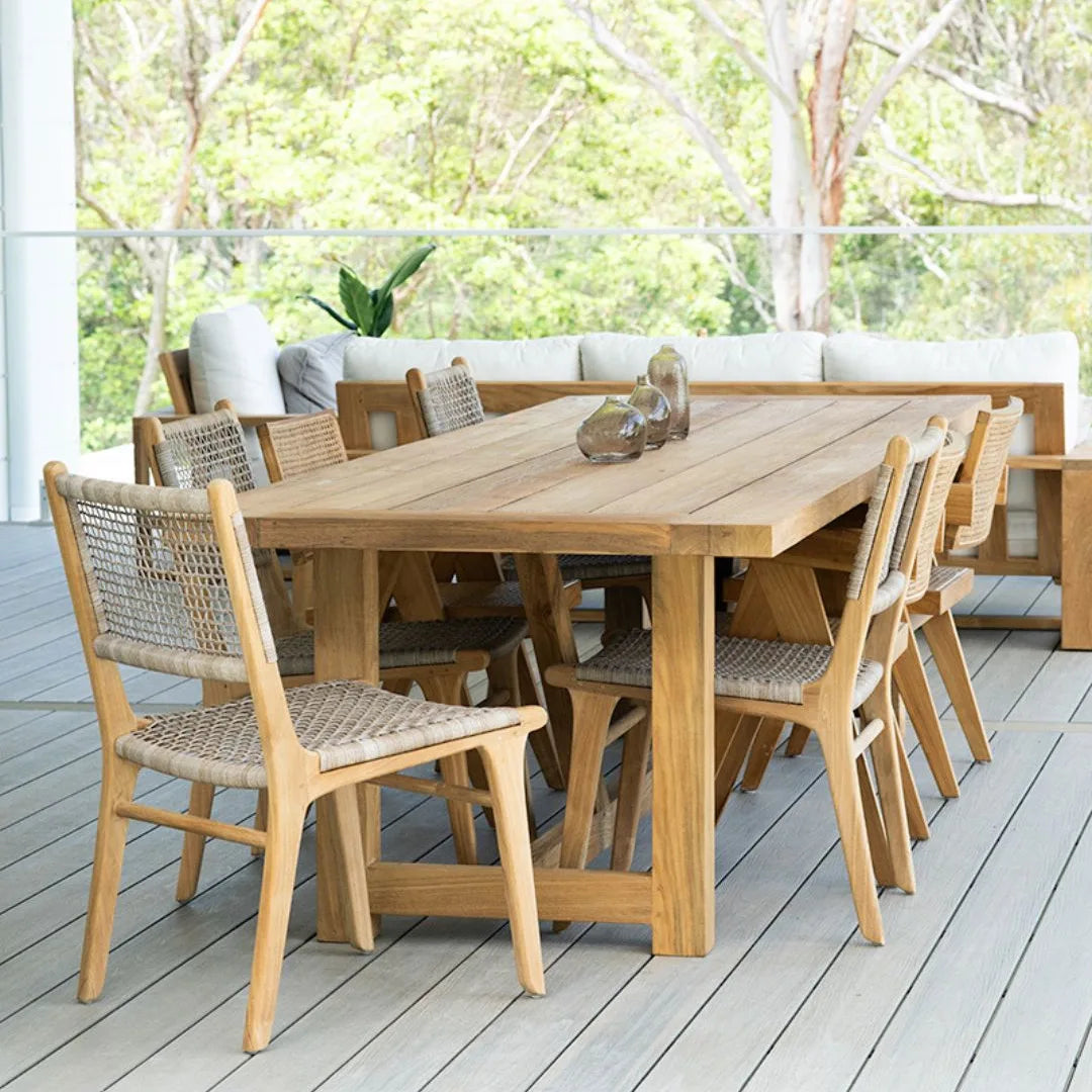 Wooden outdoor dining table with chairs on a deck with greenery in the background