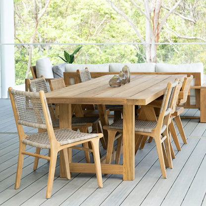 Wooden outdoor dining table with chairs on a deck with greenery in the background