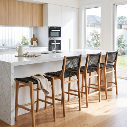 Four wooden bar stools with woven black seats and backs are arranged along a white marble kitchen island in a bright, modern kitchen.
