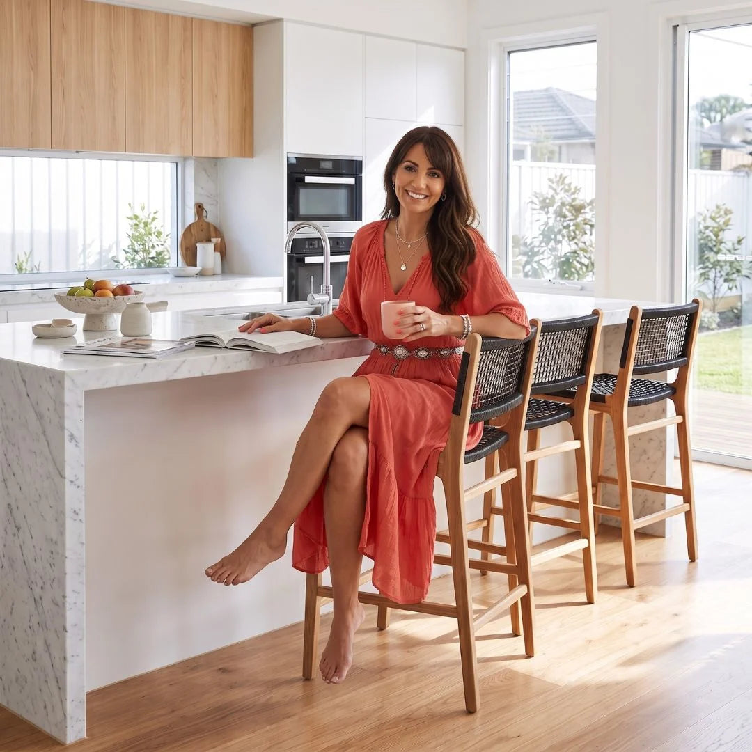 A woman in a coral-red dress sits barefoot on a wooden bar stool at a kitchen island, holding a pink mug in a bright, modern kitchen with white cabinets, wood floors, and large windows.