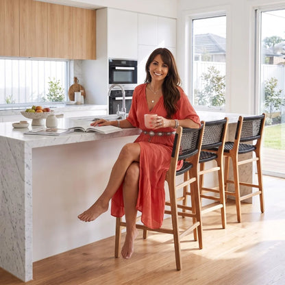 A woman in a coral-red dress sits barefoot on a wooden bar stool at a kitchen island, holding a pink mug in a bright, modern kitchen with white cabinets, wood floors, and large windows.