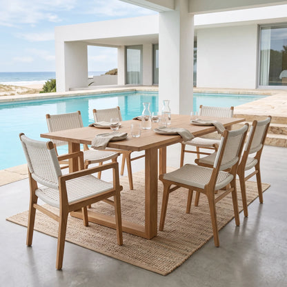 A 6-piece outdoor dining set featuring a rectangular light oak wood table and four matching woven-back chairs, styled on a jute rug next to a swimming pool with a modern house and beach in the background.