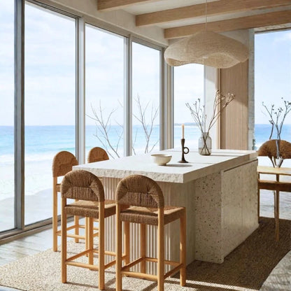 Modern kitchen with marble island and wooden chairs, large windows overlooking ocean view.