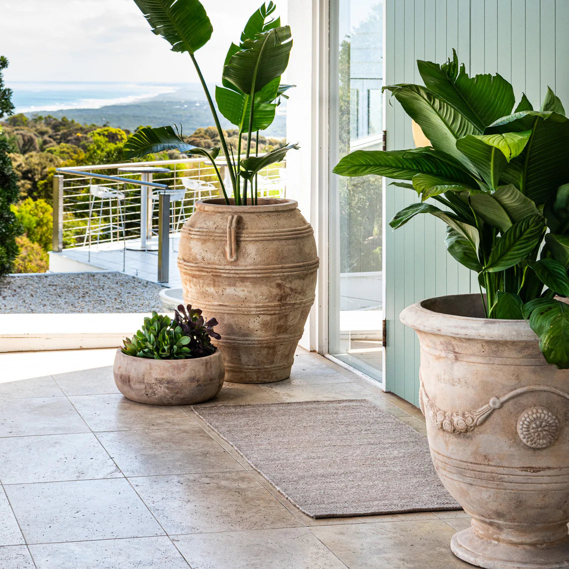 Three large decorative pots with plants on a patio with a scenic view, and a Sun Republic Eco floor mat.