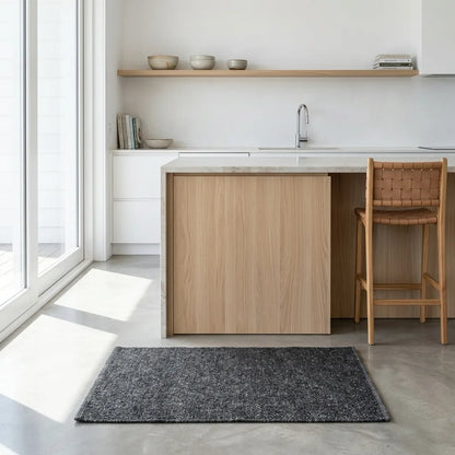 A modern kitchen island with light oak wood cabinets and a marble countertop, styled with a woven-back stool and a dark gray textured rug on a concrete floor.