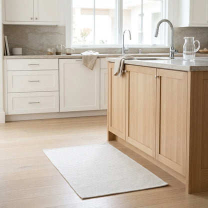 A rectangular off-white bath mat or rug placed on a light wood floor in a bright, modern kitchen with white cabinets, a wood island, and a sink.