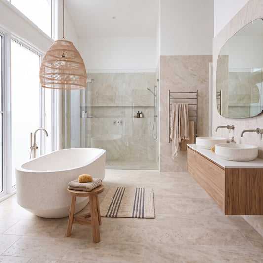 A modern bathroom interior with a freestanding white bathtub, wicker pendant light, wood stool, marble-look tile floor and walls, glass shower enclosure, double vessel sinks, and a large round mirror.