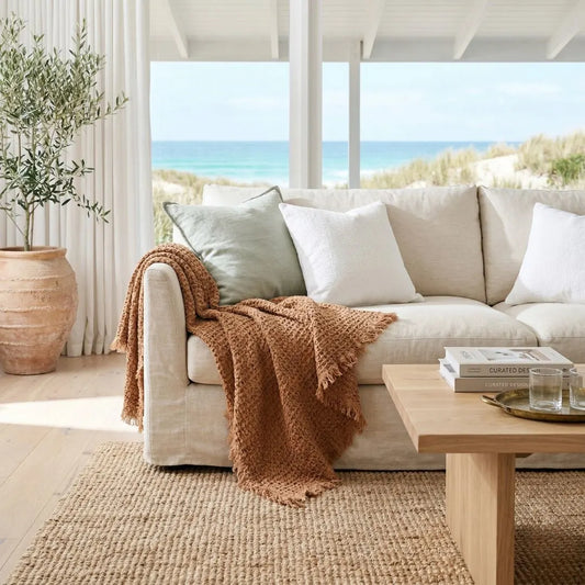 Cozy living room with a beige sofa, brown blanket, and wooden coffee table, overlooking a beach.