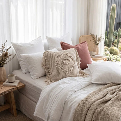 Cozy bedroom with a bed adorned with pillows and blankets, next to a window with white curtains and a cactus plant.
