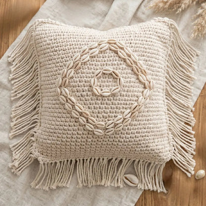 Beige woven pillow with shell design and fringes on a wooden surface