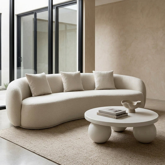 A modern curved cream sofa with four matching throw pillows in a minimalist beige living room, styled with a round white coffee table and a textured beige rug.