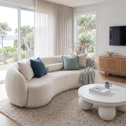 A curved cream fabric sofa in a bright, modern living room with neutral-toned decor, including blue and sage green throw pillows, a round white coffee table, and large windows overlooking greenery.