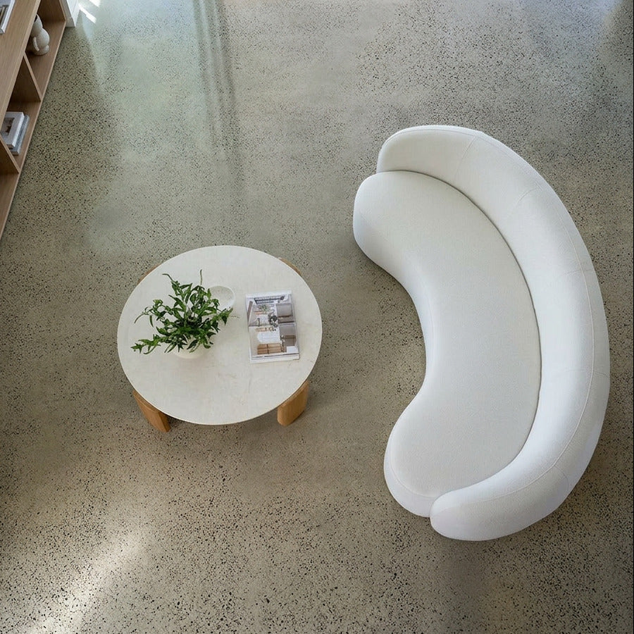A modern curved white lounge chair next to a round white coffee table with a small green plant and magazine, set on a polished concrete floor.