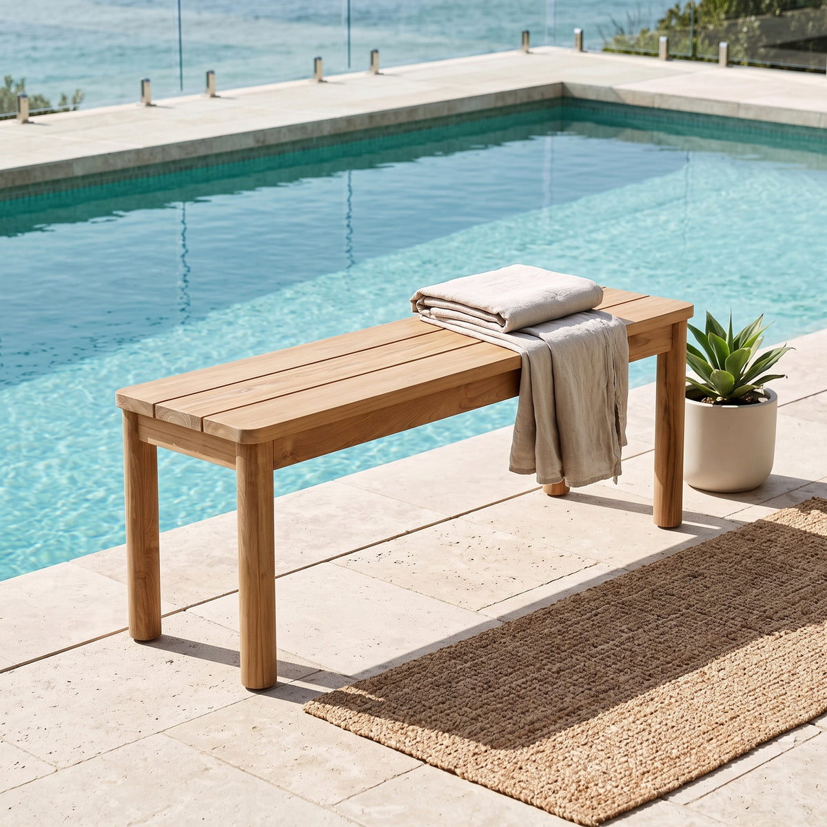 A light natural wood outdoor bench sits beside a swimming pool on a tiled patio, with folded beige textiles draped over the back and a small potted succulent nearby.