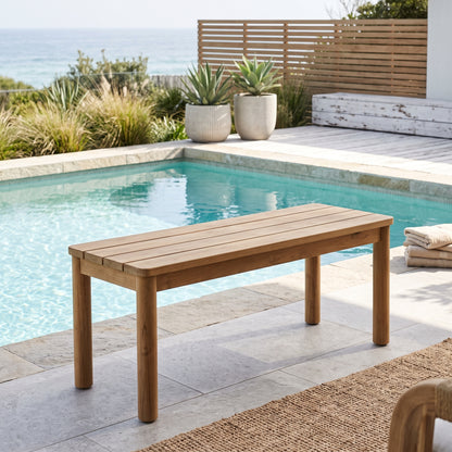 A rectangular outdoor wooden bench with a slatted seat and four legs, positioned beside a swimming pool on a stone patio with a poolside view and plants in the background.