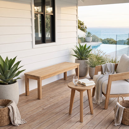 A styled outdoor patio scene featuring a light natural wood bench, a small round wooden side table with an open book and glass, and a light-colored armchair with a draped throw blanket. The furniture is arranged on a wooden deck next to large potted green plants, with a white house wall and an ocean view in the background.