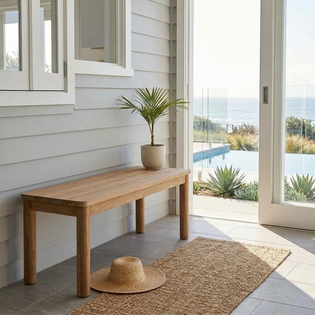 A natural wood entryway bench with a potted palm and a straw hat, set on a tiled porch with a jute rug and a view of the ocean through glass doors.