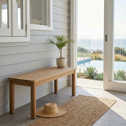 A natural wood entryway bench with a potted palm and a straw hat, set on a tiled porch with a jute rug and a view of the ocean through glass doors.