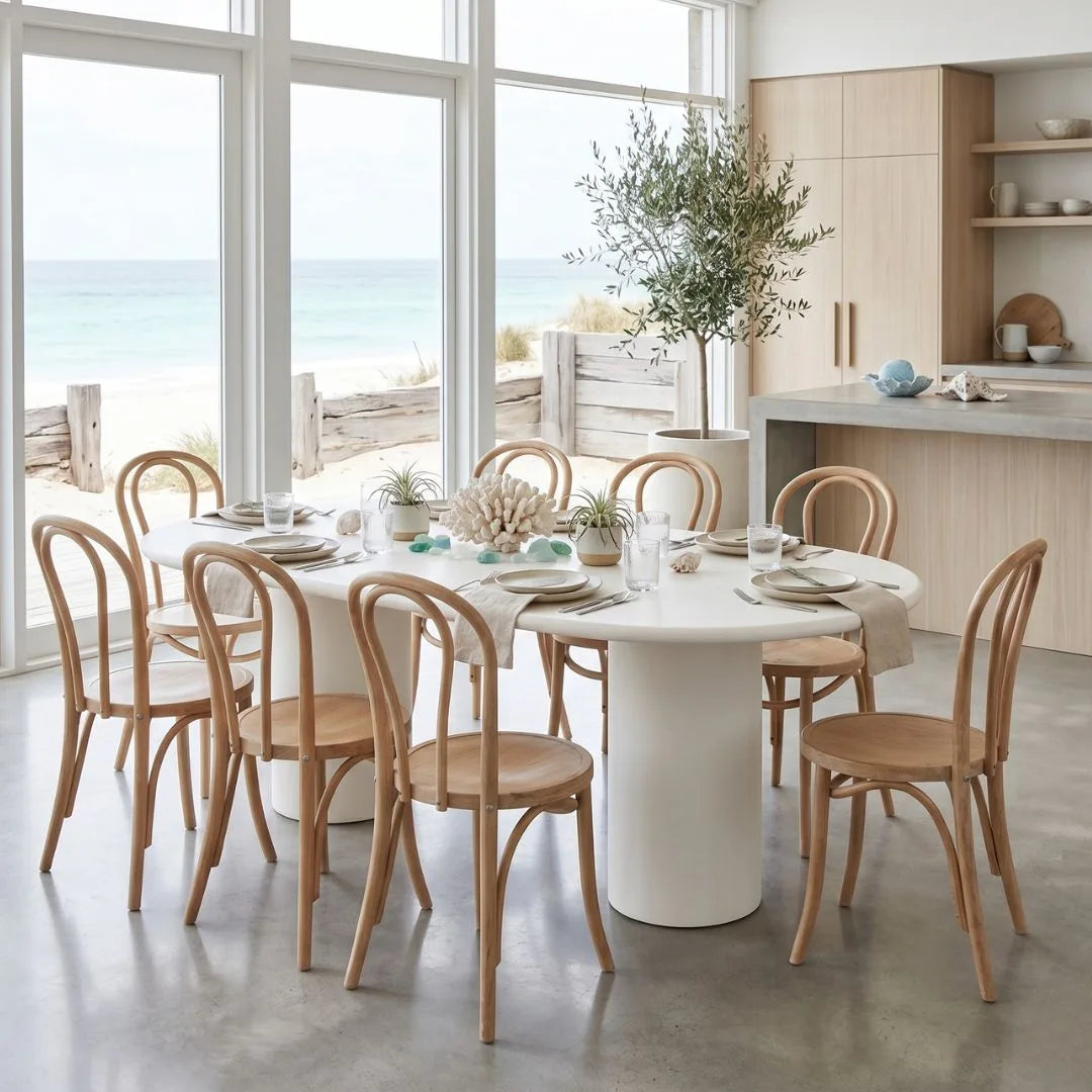 Dining area with a round white table and wooden chairs in a bright room with large windows.