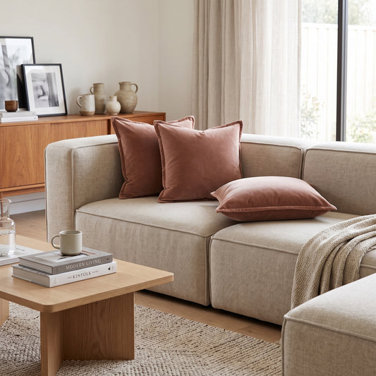 A modern beige sectional sofa in a bright, contemporary living room, styled with three dusty rose velvet throw pillows and a beige knit throw blanket.