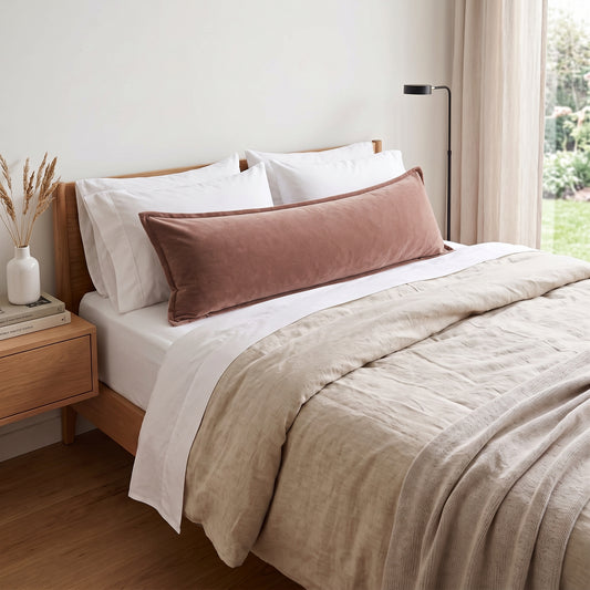 A bedroom scene showing a bed styled with white pillows, a dusty rose velvet lumbar pillow, a beige linen duvet, and a wooden bed frame. A window with sheer curtains and a black floor lamp are visible in the background.