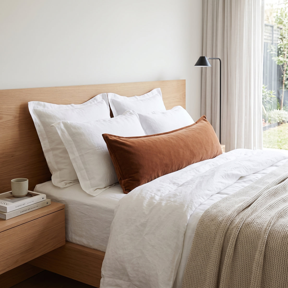 A styled bedroom scene showing a bed with white bedding, multiple white pillows, and a prominent rust-brown lumbar pillow. The bed has a light wood headboard and matching nightstand, with a black floor lamp and a window in the background.