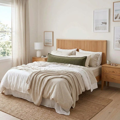 A neatly made bed in a bright, neutral-toned bedroom, featuring a light wood slatted headboard, beige and off-white bedding, a green accent pillow, matching nightstands, and a woven rug.