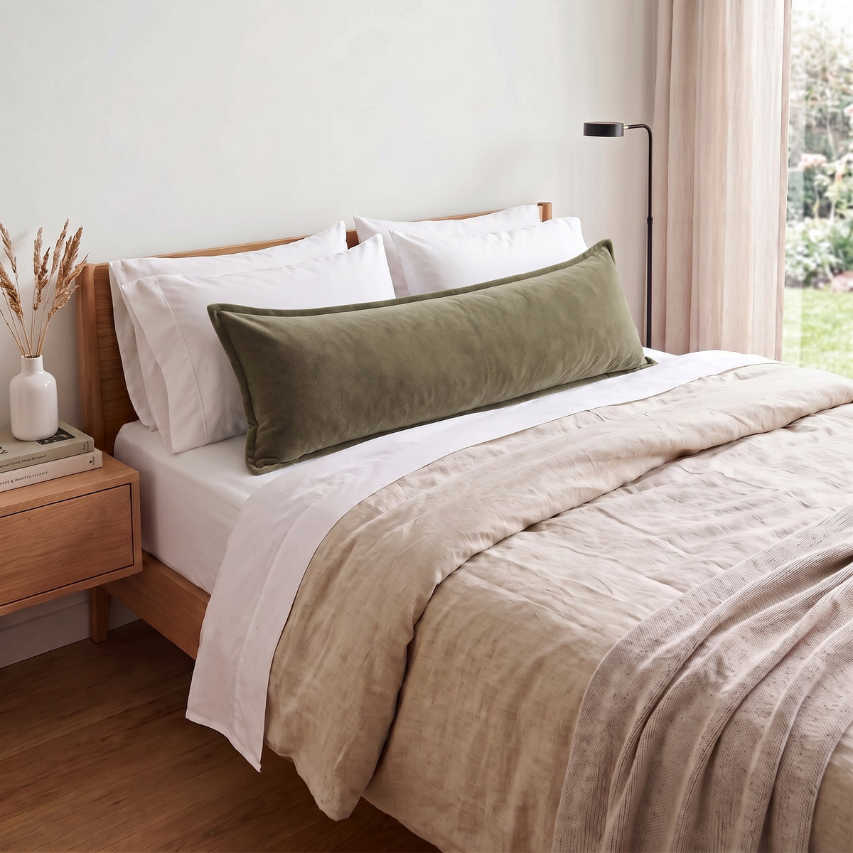 An olive green velvet lumbar pillow resting on a bed with white pillows and a beige linen duvet, set in a softly lit bedroom.