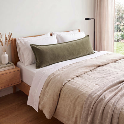 An olive green velvet lumbar pillow resting on a bed with white pillows and a beige linen duvet, set in a softly lit bedroom.