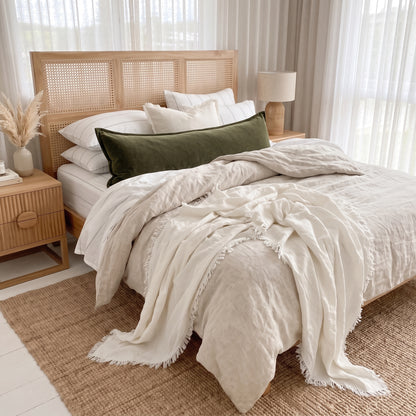 A styled bed in a bright, neutral-toned bedroom featuring a cream linen duvet cover and throw blanket with fringe, white pillows, a dark olive green velvet lumbar pillow, and a light wood rattan headboard.