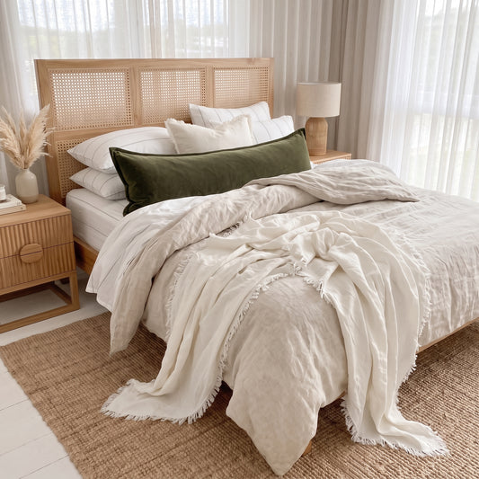 A styled bed in a bright, neutral-toned bedroom featuring a cream linen duvet cover and throw blanket with fringe, white pillows, a dark olive green velvet lumbar pillow, and a light wood rattan headboard.