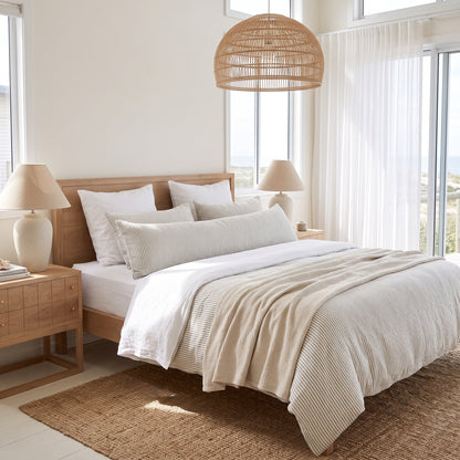 A neatly made bed in a bright, modern bedroom featuring a light wood headboard and footboard, white bedding, textured cream duvet, striped pillows, matching bedside tables, table lamps, a woven pendant light, and a large window with sheer curtains.