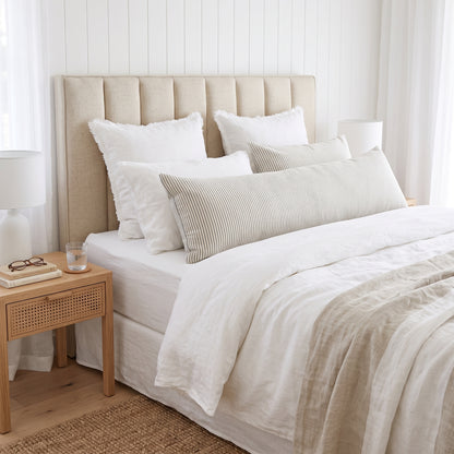 A neatly made bed in a bright bedroom setting, featuring a tall upholstered headboard, layered white and beige bedding, striped lumbar pillows, a wooden bedside table with a lamp, and a woven rug.