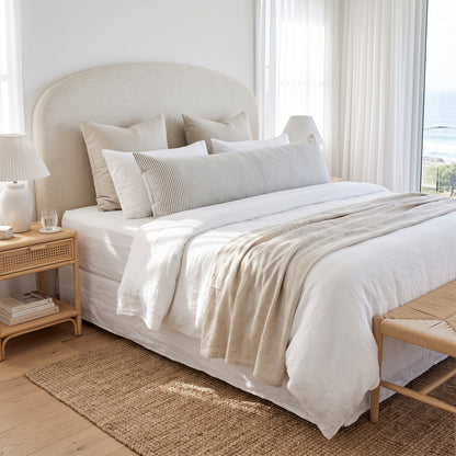 A neatly made bed in a bright, minimalist bedroom featuring a cream upholstered headboard, white bedding, neutral beige linens, striped pillows, and a textured throw blanket. A light wood nightstand with a white lamp sits on the left, and a woven rug covers the wooden floor.