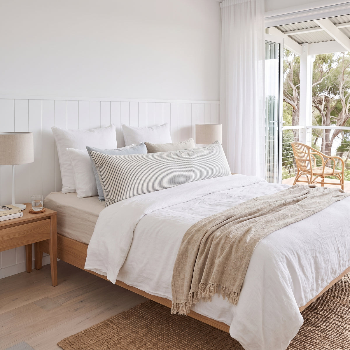 A neatly made bed in a bright, modern bedroom with white bedding, a beige throw blanket, a wooden bed frame, a bedside table with a lamp, and large windows looking out to a balcony with a chair.