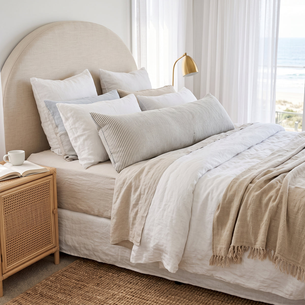 A neatly made bed in a bright bedroom setting, featuring a beige upholstered headboard, layered white and beige linens, a striped lumbar pillow, and a fringed beige throw blanket. A light wood nightstand with a woven cane front sits beside the bed, and sheer curtains frame a window in the background.