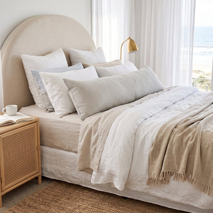 A neatly made bed in a bright bedroom setting, featuring a beige upholstered headboard, layered white and beige linens, a striped lumbar pillow, and a fringed beige throw blanket. A light wood nightstand with a woven cane front sits beside the bed, and sheer curtains frame a window in the background.