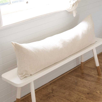 A long cream-colored textured lumbar pillow resting on a white bench with light wood legs in a bright room with white paneled walls and a wood floor.