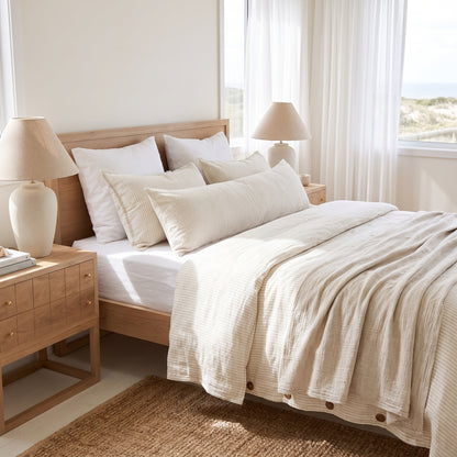 A neatly made bed in a bright, sunlit bedroom featuring cream-colored textured cotton bedding, including a duvet cover and matching pillows, styled with a light wood bed frame, bedside tables with lamps, and a window looking out to the sea.