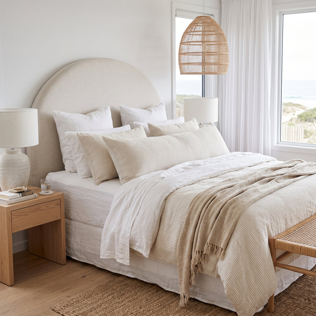 A beautifully styled beach-inspired bedroom featuring a bed dressed in cream and beige textured bedding, a large round upholstered headboard, a wooden nightstand, and a rattan pendant light.