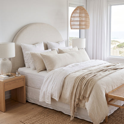 A beautifully styled beach-inspired bedroom featuring a bed dressed in cream and beige textured bedding, a large round upholstered headboard, a wooden nightstand, and a rattan pendant light.