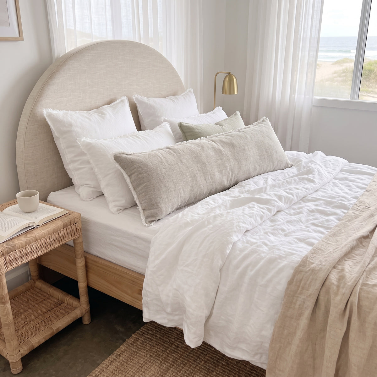 A neatly made bed in a bright bedroom with a large upholstered headboard, white bedding, and several light beige pillows. A woven side table, floor lamp, and window with sheer curtains are visible, with a beach scene outside.