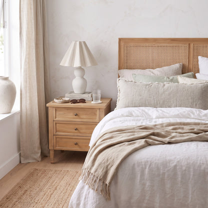 A styled bedroom scene featuring a light wood bed frame with a woven cane headboard, dressed in white linens and a beige throw blanket, next to a matching wooden nightstand with a white table lamp.