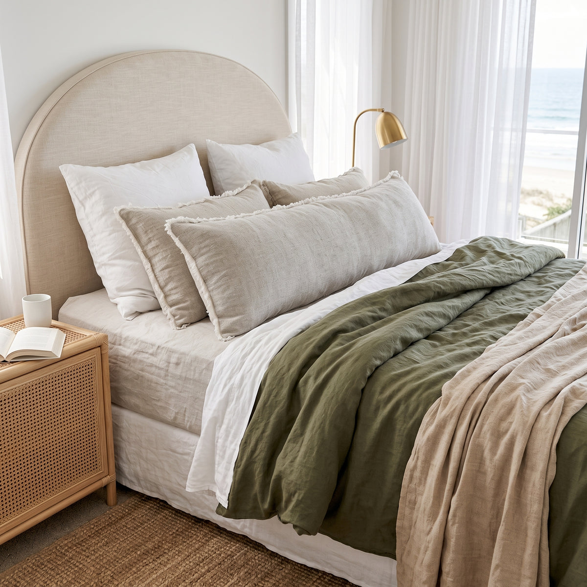 A neatly made bed in a bright bedroom setting, featuring a beige upholstered headboard, layered white and beige pillows, an olive green duvet, and a tan throw blanket. A woven nightstand, gold bedside lamp, and large window with ocean view are visible.