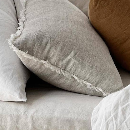 A close-up of a light gray linen throw pillow with frayed edges, resting on a matching gray linen bedspread alongside other pillows.