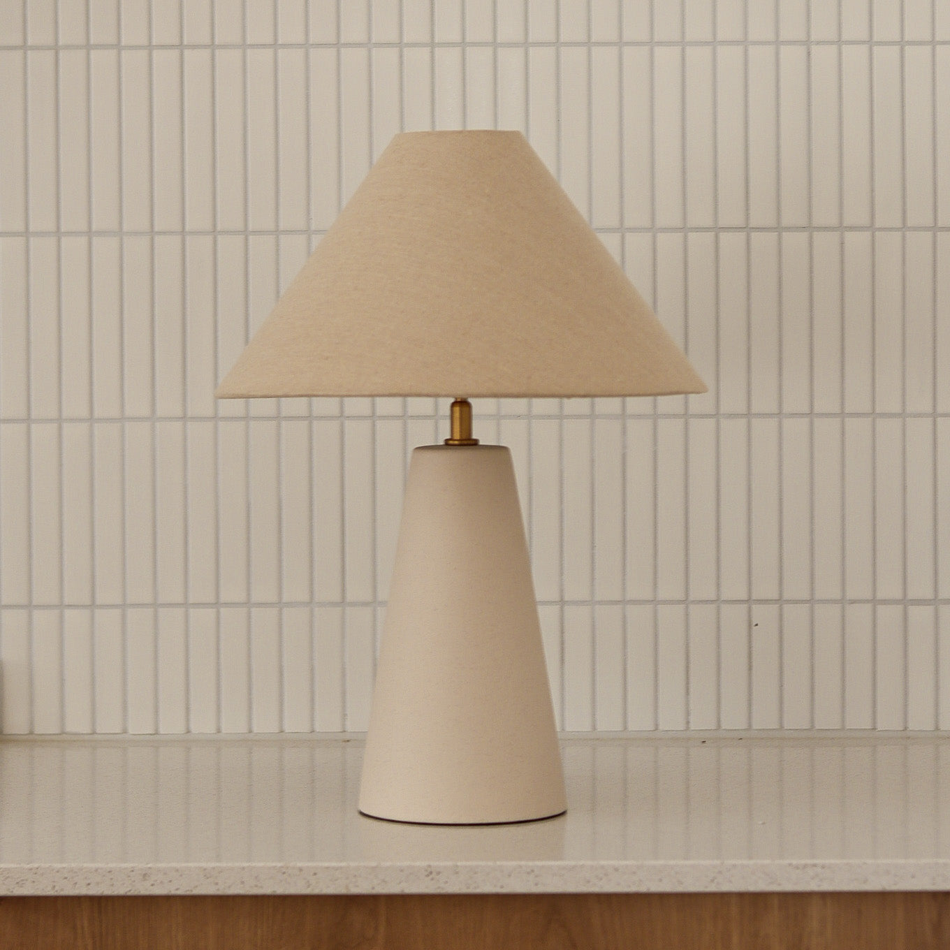 Beige table lamp with a cone-shaped base against a tiled wall.
