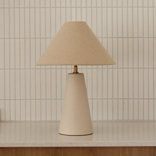 Beige table lamp with a cone-shaped base against a tiled wall.