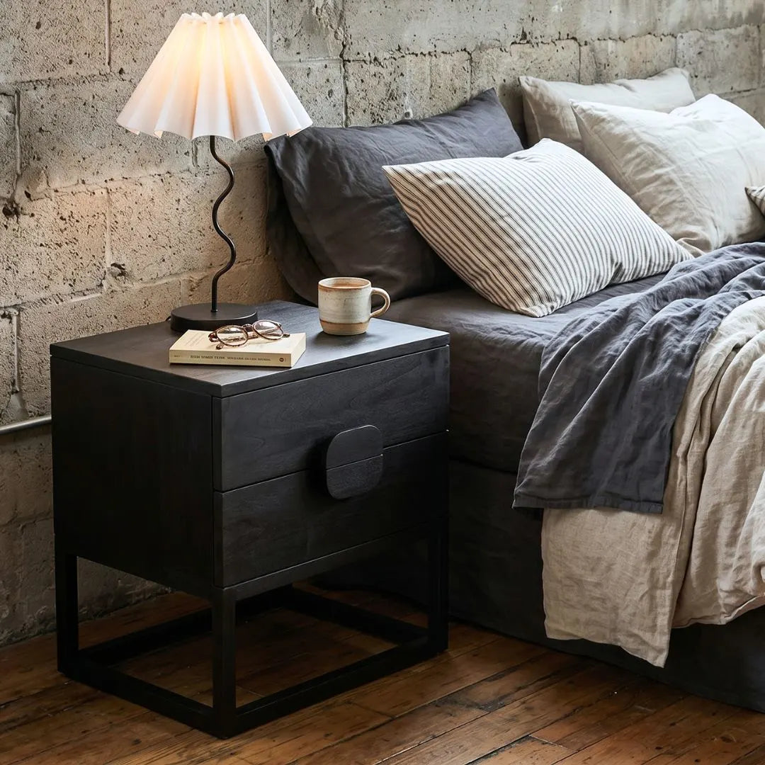 Black nightstand with lamp and mug next to a bed with pillows and blankets against a stone wall.