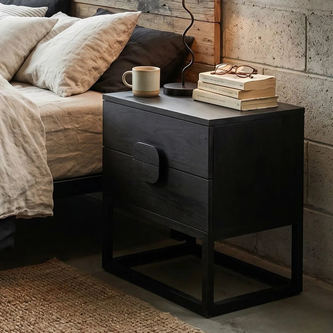 Black nightstand with a mug, books, and glasses next to a bed with pillows.